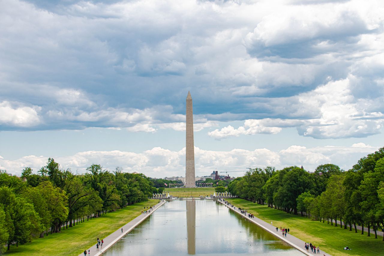 gallery-4 A scenic view of the Washington Monument in Washington, D.C., with reflections in the reflecting pool and a cloudy sky overhead.